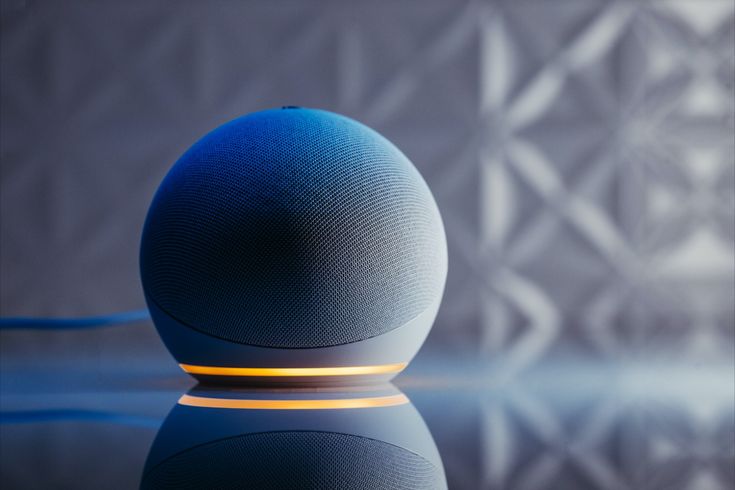 Imagem em close-up do Echo Dot de última geração na cor branca, posicionado sobre uma superfície reflexiva. A luz inferior do dispositivo está acesa em um tom quente, criando um contraste com a luz ambiente mais fria. O fundo apresenta uma parede texturizada em relevo, focando a atenção no design elegante do smart speaker.